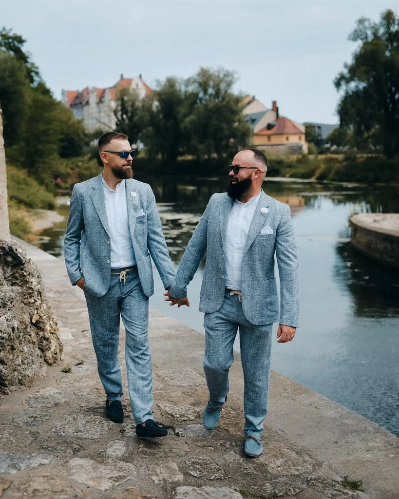 Zwei Bräutigame in hellblauen Anzügen spazieren Hand in Hand am Ufer bei der Steinernen Brücke in Regensburg