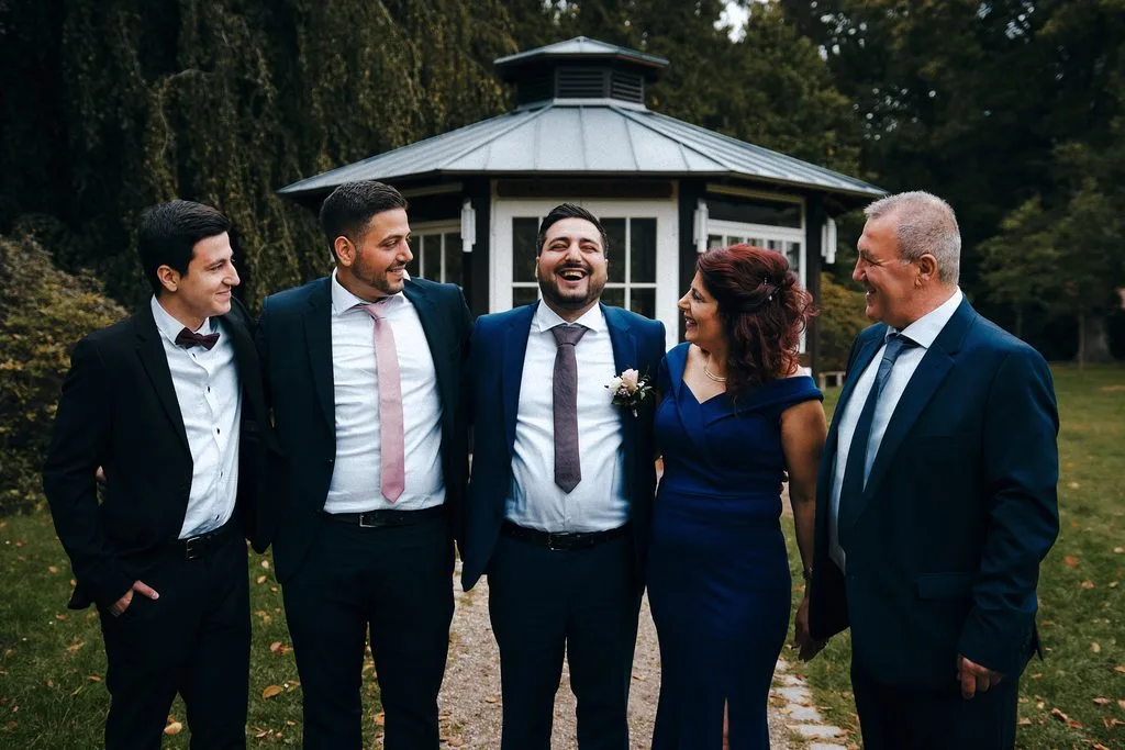 Lachender Bräutigam mit Familie und Freunden vor einem Gartenpavillon