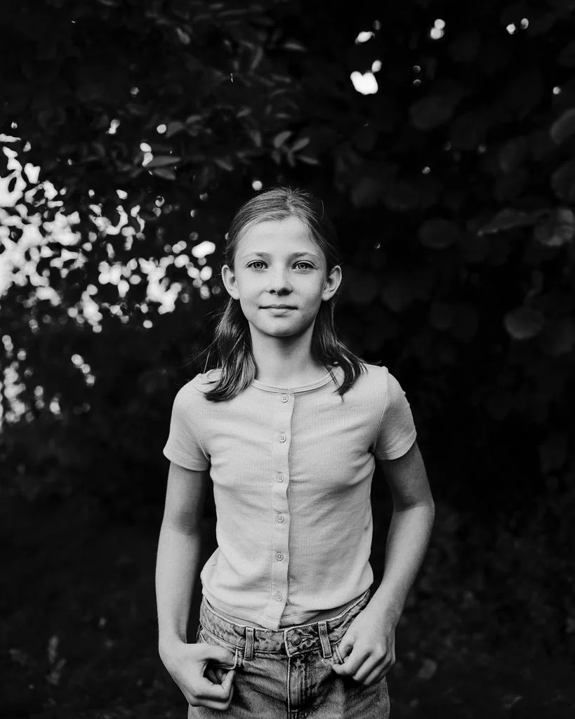 Schwarz-Weiß-Portrait eines Mädchens mit den Händen in den Hosentaschen vor dunklem Laub