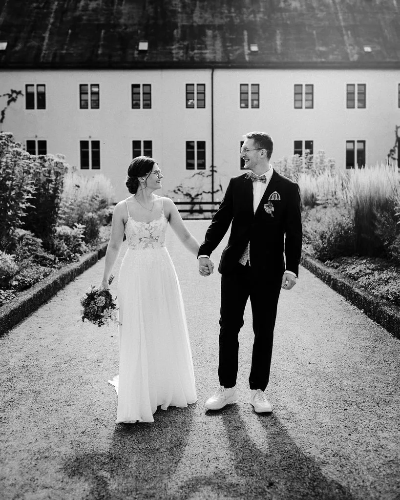 Brautpaar geht Hand in Hand durch einen Garten vor einem historischen Gebäude, Schwarz-Weiß