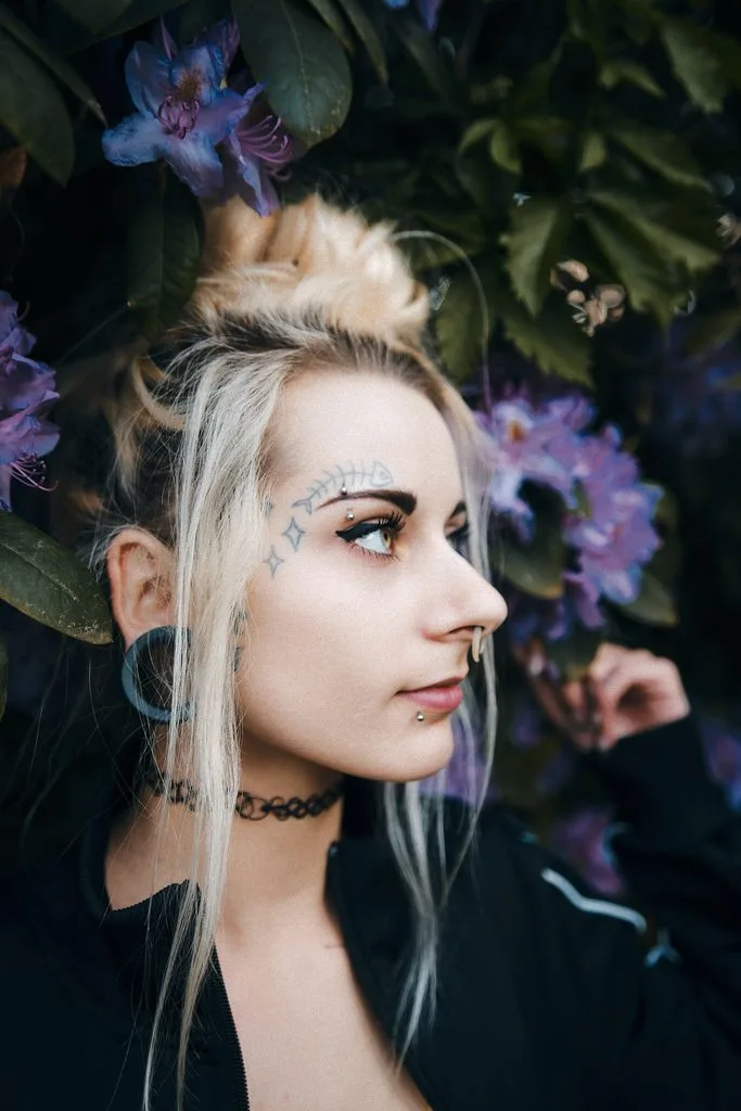 Portrait einer Frau mit blondem Dutt und Gesichtstattoos vor blühendem Rhododendron