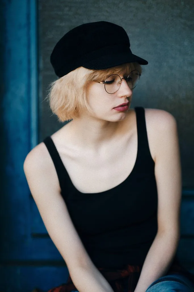 Portrait einer jungen Frau mit Brille und Schiebermütze vor einer blauen Wand