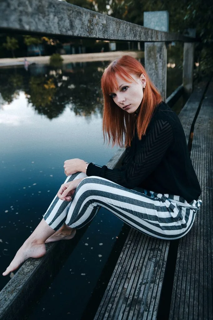 Frau mit roten Haaren und gestreifter Hose sitzt barfuß auf einem Steg über dem Wasser
