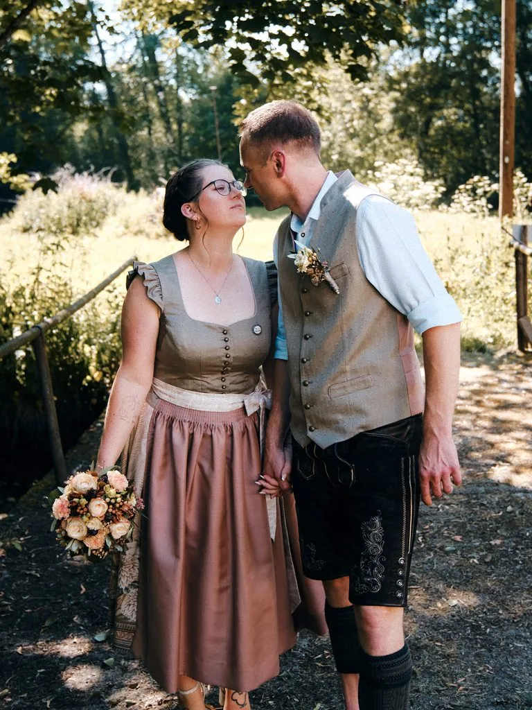 Brautpaar in Dirndl und Lederhose küsst sich auf einem sonnigen Waldweg