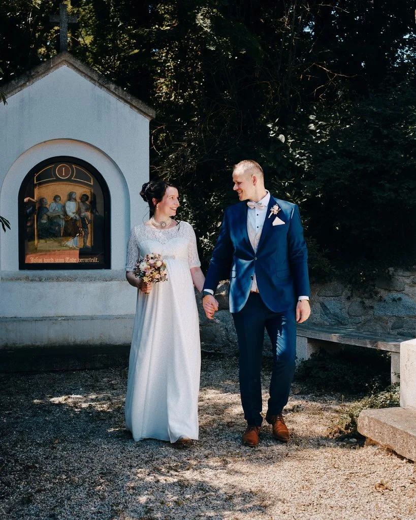 Brautpaar spaziert Hand in Hand an einer Wegkapelle vorbei