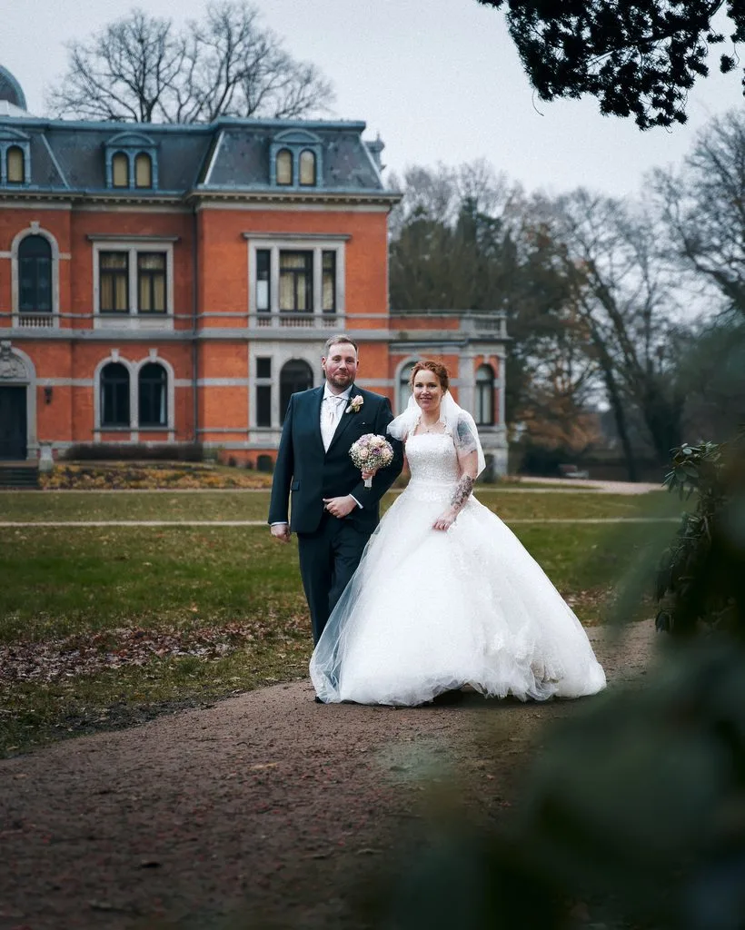 Brautpaar spaziert vor einer historischen Backsteinvilla durch einen herbstlichen Park